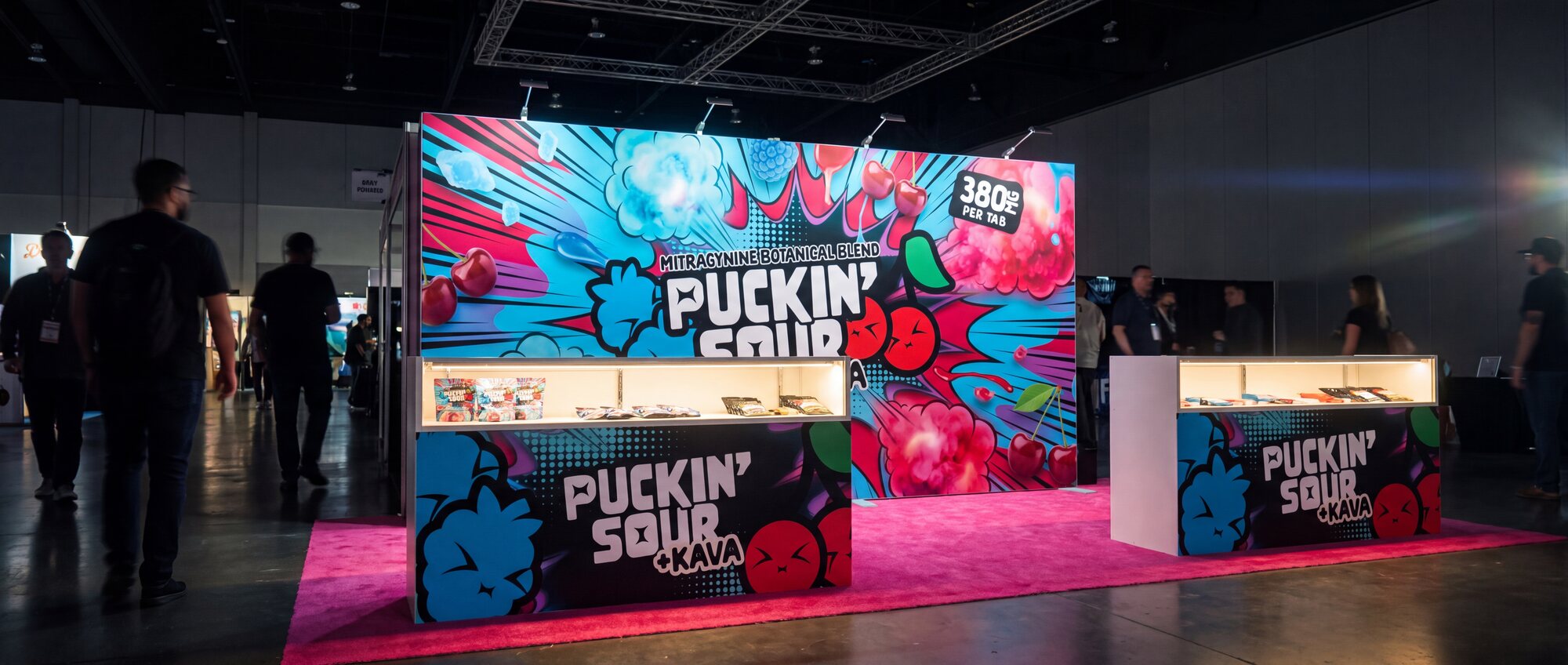 FIG.09 — the Puckin' Sour +KAVA booth glowing in a dim hall at Champs Las Vegas, matching counters lit from within on hot pink carpet